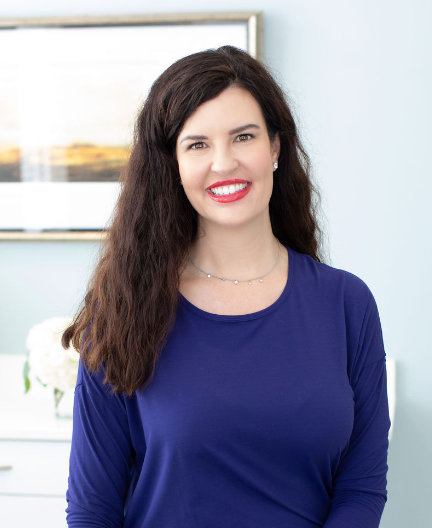 A woman with long dark hair is smiling, wearing a blue top, standing indoors with a blurred framed picture and flowers in the background.