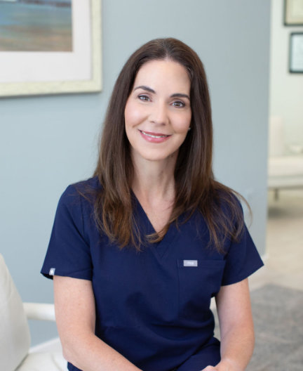 A woman with long brown hair, wearing navy blue scrubs, sits in a bright room with framed certificates on the wall.