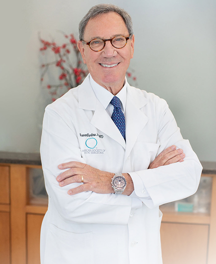 A doctor in a white coat stands with arms crossed. He wears glasses and a blue tie, with a name badge visible. Background includes a wooden counter and red decorative branches.