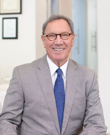 Man in a gray suit and blue tie, wearing round glasses, smiling while seated. Background shows framed certificates and a door.
