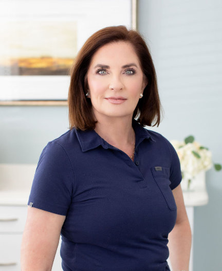 A woman with brown hair wearing a navy polo shirt stands indoors, looking at the camera. A framed picture and flowers are blurred in the background.