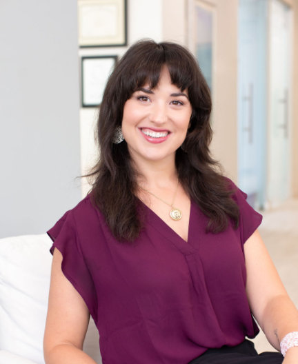 A woman with shoulder-length dark hair, wearing a purple blouse, sits indoors smiling at the camera.