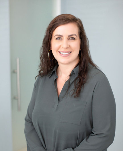 A woman with long wavy hair smiles, wearing a dark green blouse and hoop earrings. She is standing indoors against a light-colored wall with a door handle visible in the background.