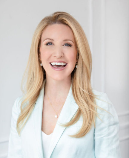 Smiling person with long blonde hair wearing a light-colored blazer and a white top, standing against a plain white background.