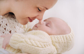 An adult gently touches noses with a sleeping baby dressed in a knitted cream outfit.