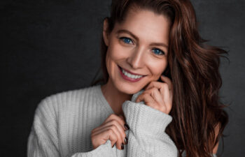 A headshot of a warmly smiling woman with long brown hair and striking blue eyes, wearing a cozy white sweater against a dark gray background.